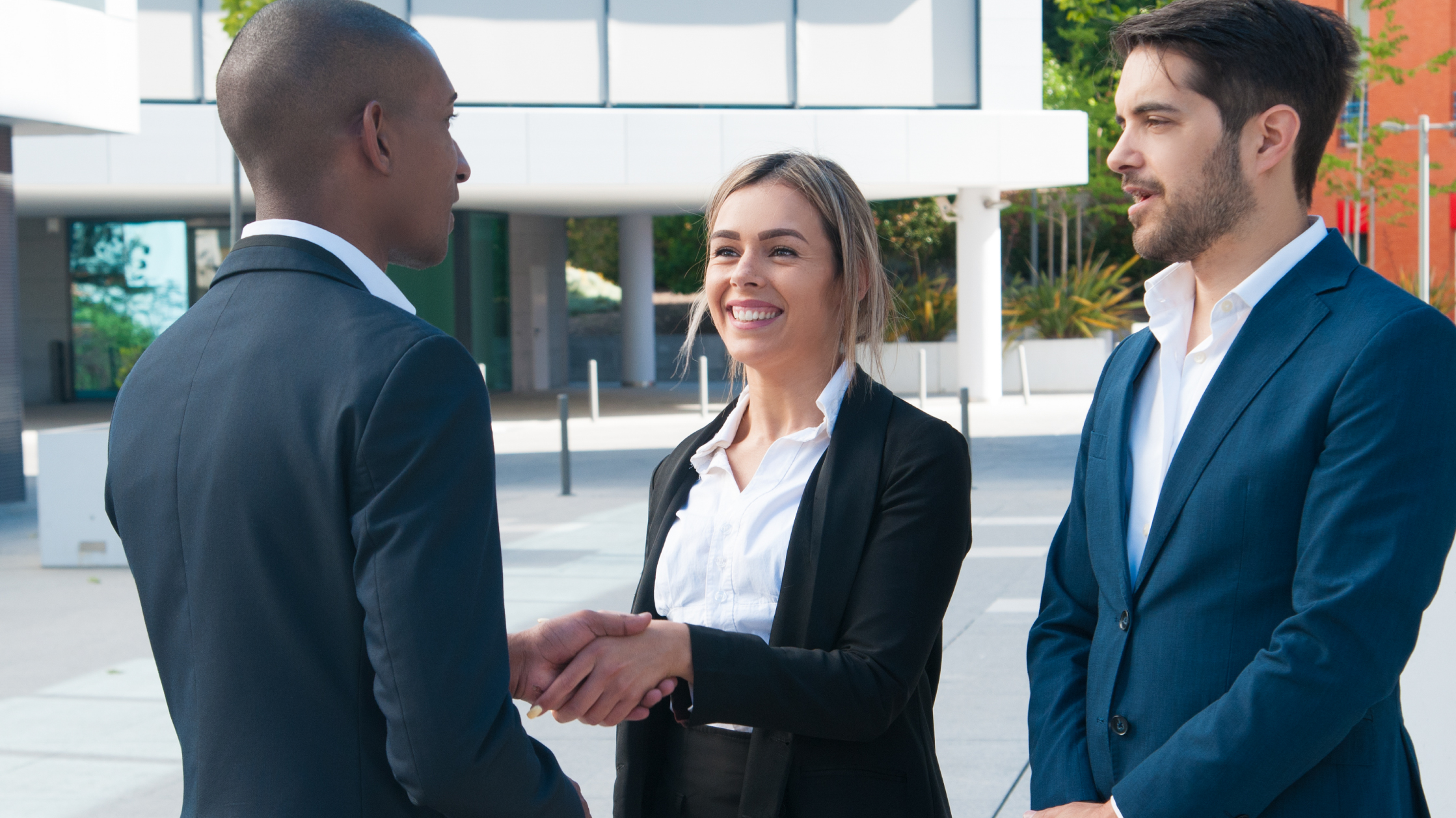 A smiling woman shakes the hand of a man while another man watches beside her. They are all wearing suits and are outside with a white building behind them.