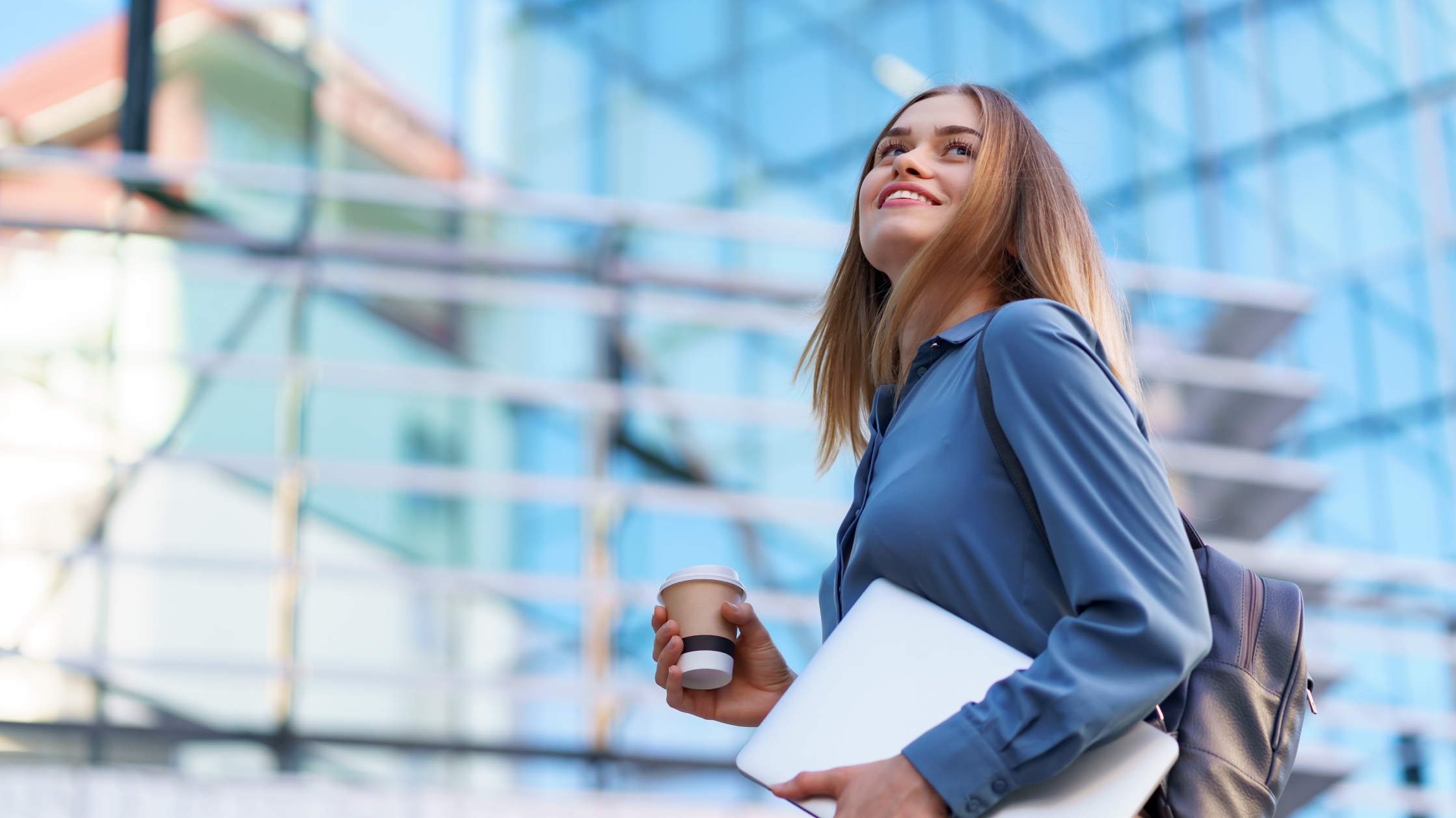 Une femme souriante portant une chemise bleue et tenant une tasse de café et un ordinateur portable. Derrière elle, un bâtiment en verre flou.