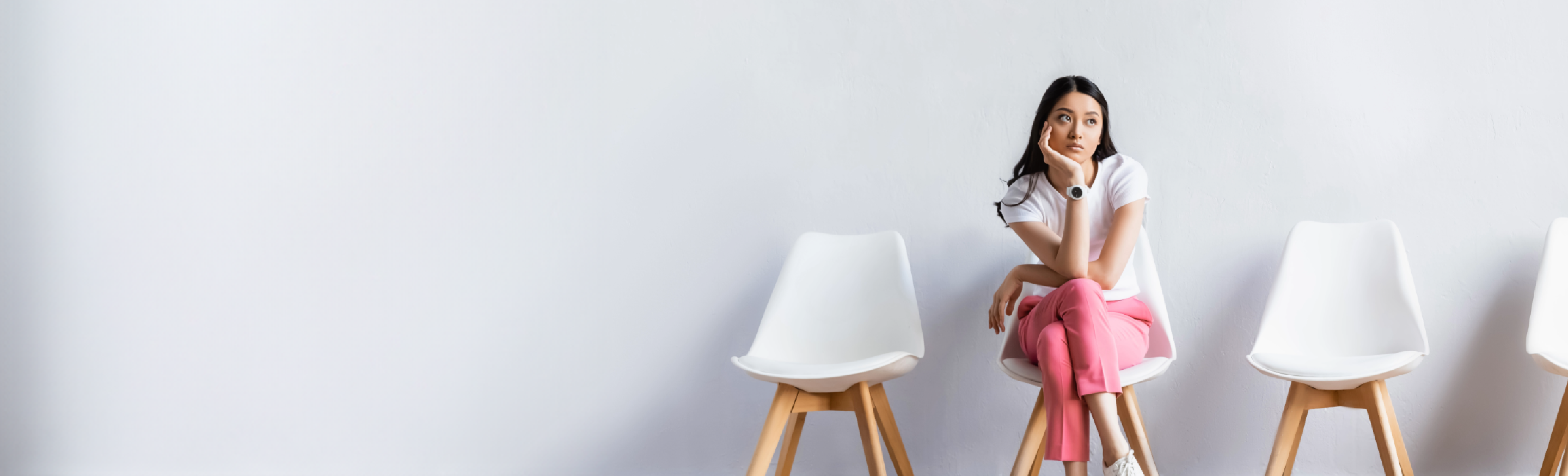 A woman in a white shirt and pink pants, sits alone on one of four white chairs with a bored expression.