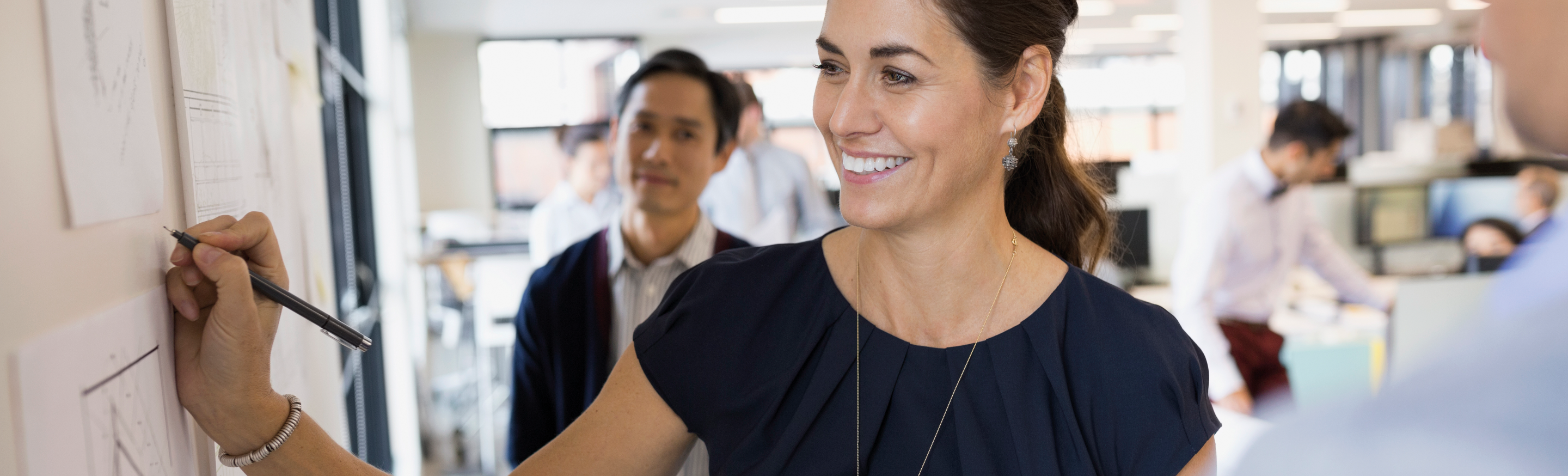 A woman in a dark blue blouse smiles brightly as she writes on a whiteboard while a man looks on behind her in an office setting.