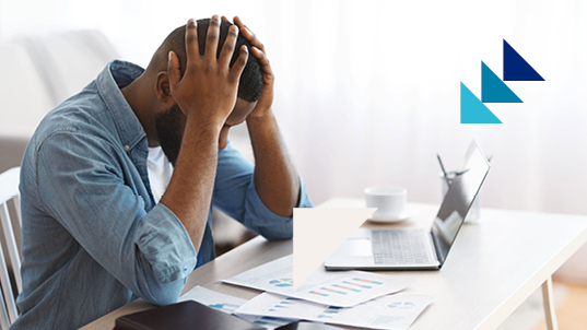 A man on a desk looks down on report pages with his hands holding his head, overwhelmed. On the desk sits a laptop and a cup of coffee.