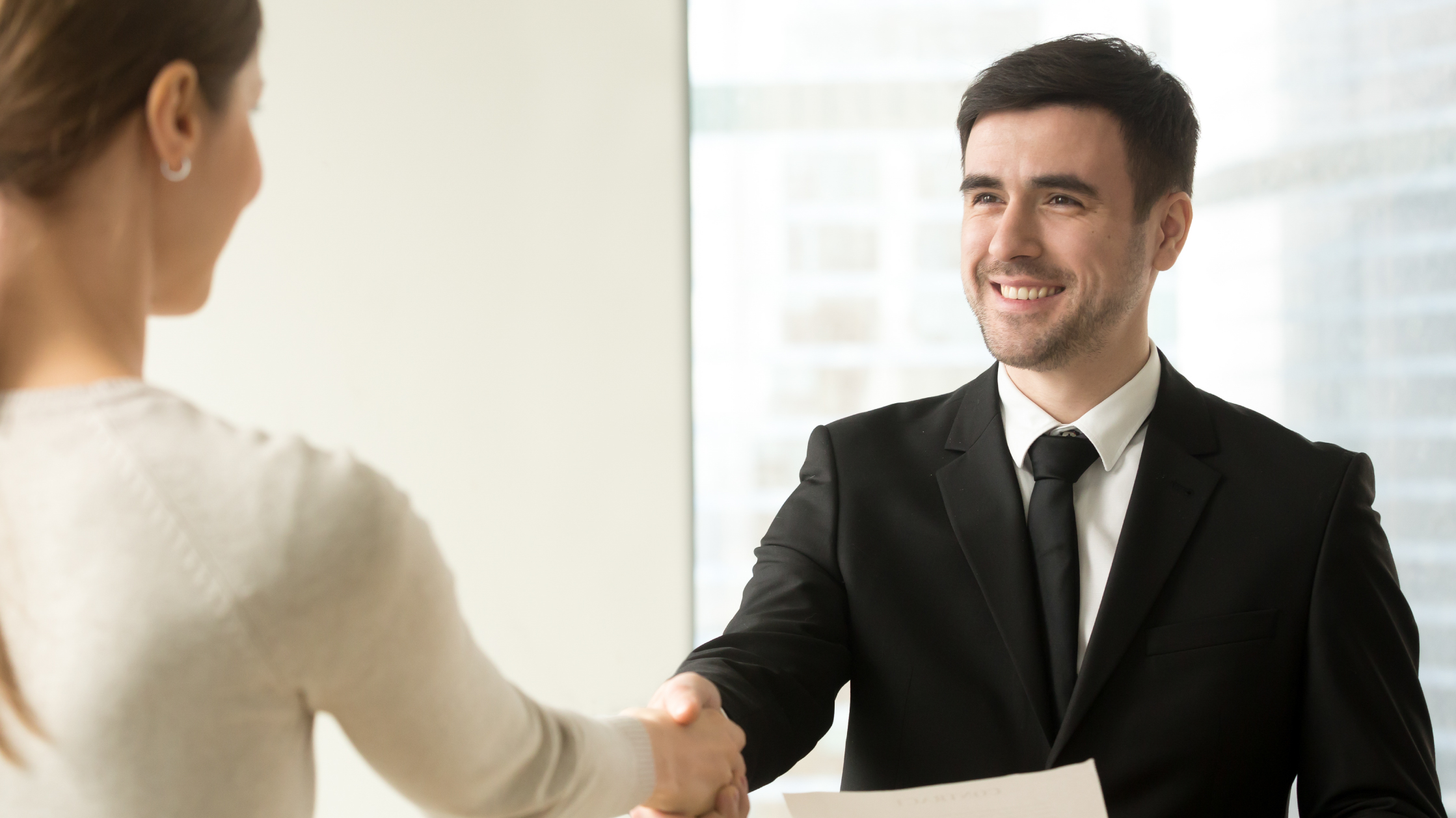 A man in a black suit and tie smiles as he shakes hands with a woman.