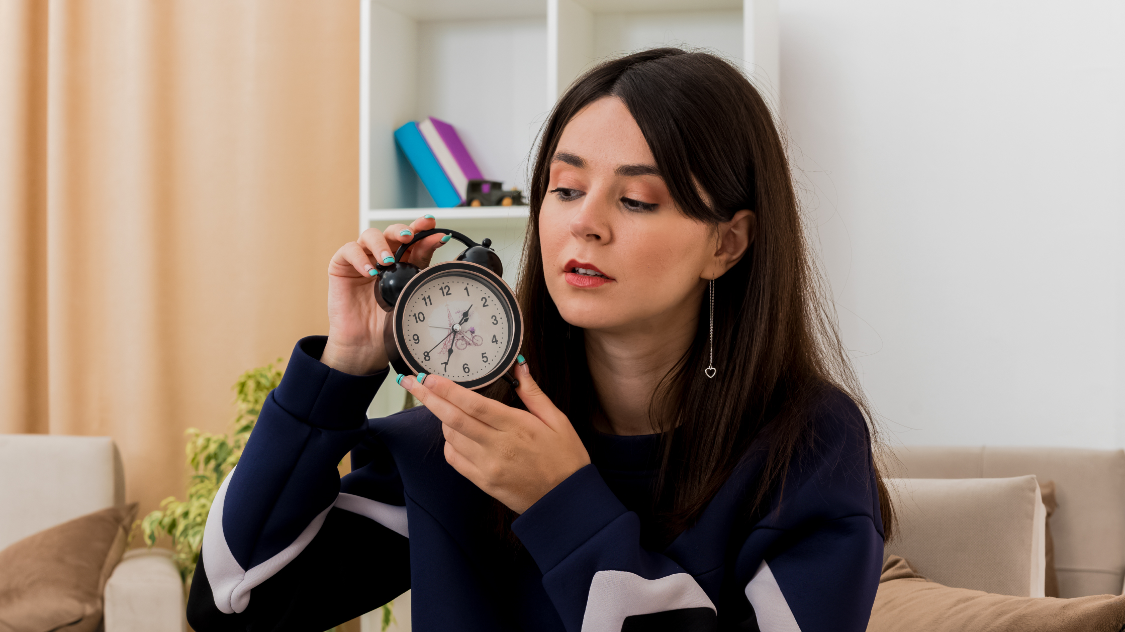 A woman holds an alarm clock and looks at it. She is in her living room with a beige sofa behind her and also a white bookshelf.