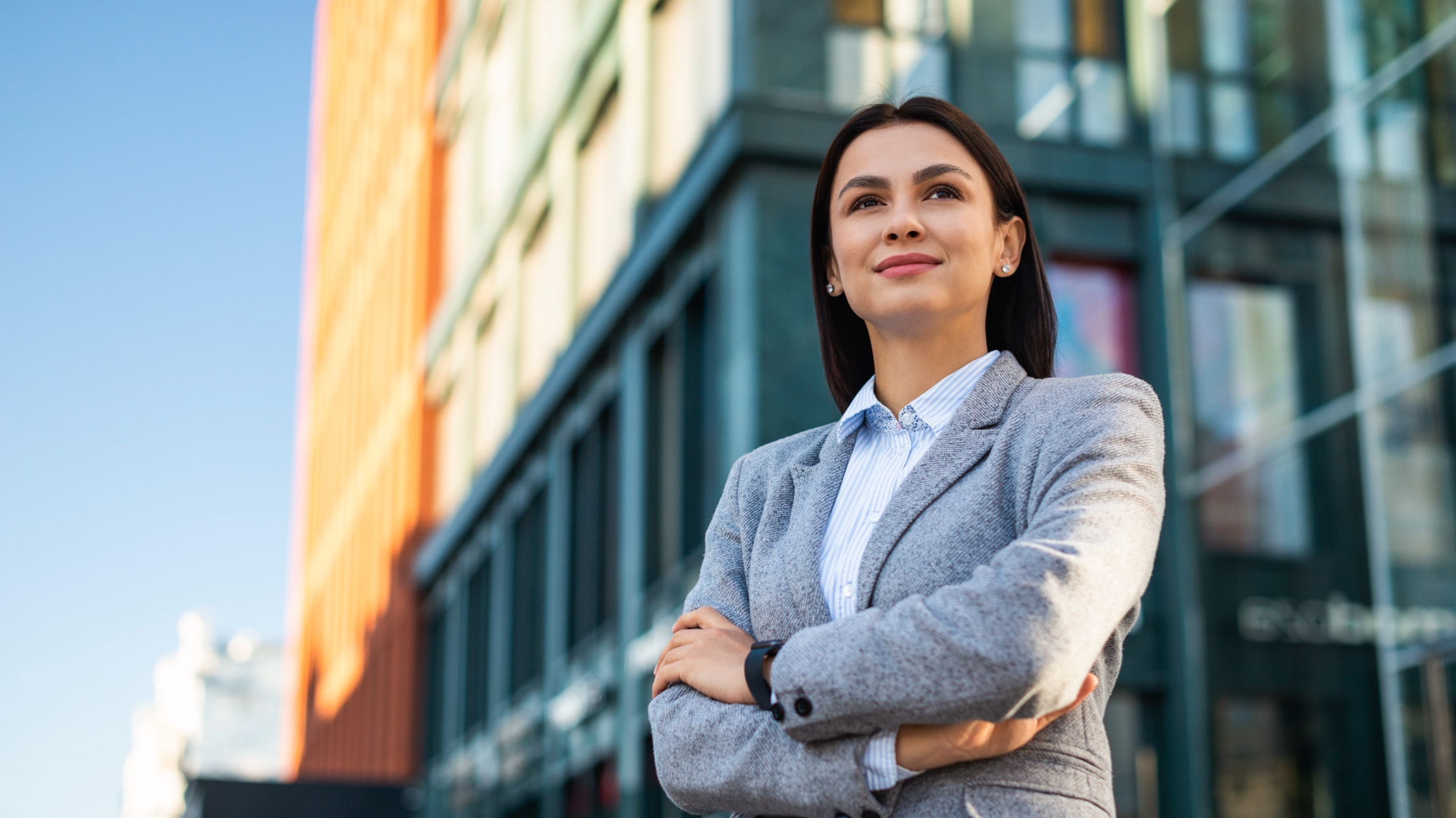 A woman looks into the distance with a pleased smile on her face. Her arms are crossed and she's wearing a gray suit with a striped blue and white shirt. Behind her are several buildings.