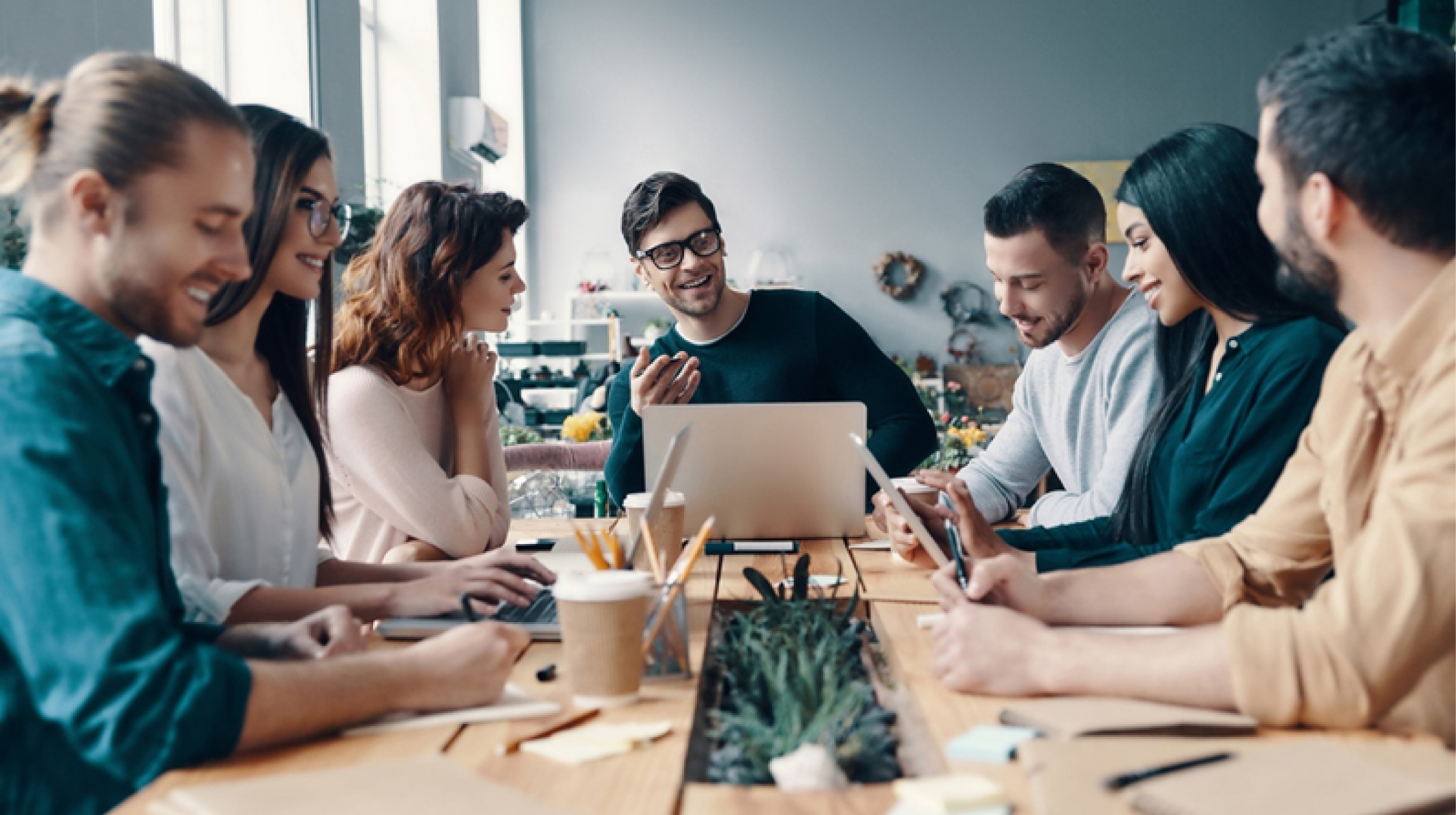 A group of men and woman smile while on a meeting. In the center the leader is a smiling man with glasses and a green sweater. On the table they sit at are several coffee cups, pencils, pads and a laptop.