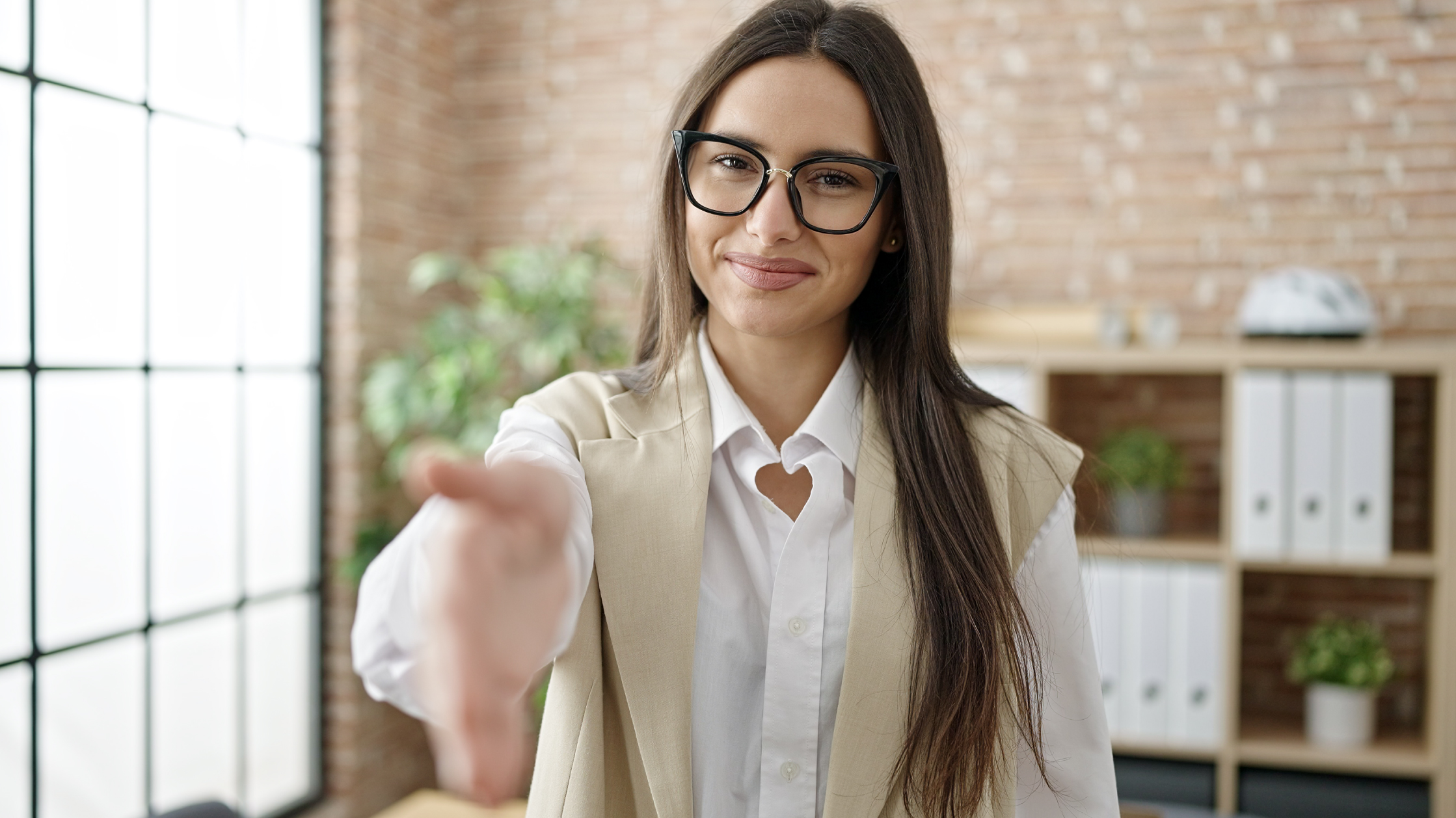 A woman holds her hand out with a smile on her face. She wears thin-rimmed black glasses and a beige vest over a white dress shirt. She is standing in an office with brick walls.