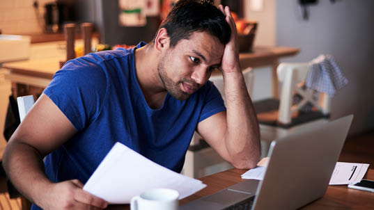 A man wearing a blue shirt holds his head and looks into his laptop with a troubled look.